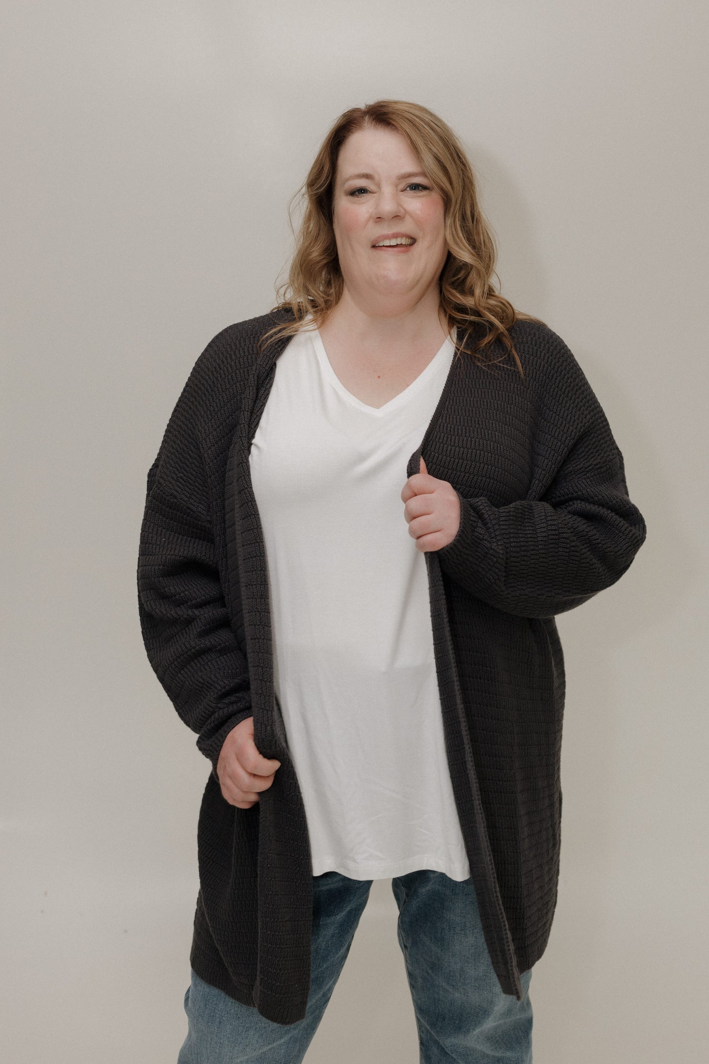 Person wearing a black cardigan over a white shirt and blue jeans on a plain background