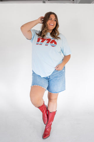 Woman wearing a light blue t-shirt with '1776' printed on it, denim shorts, and red cowboy boots against a white background.