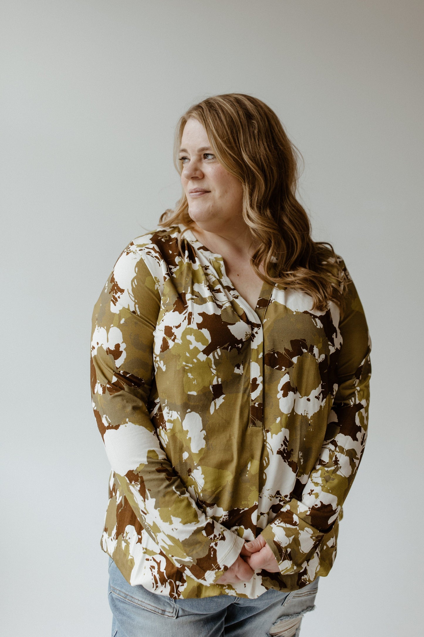 Against a plain background, a person with long hair is wearing the Yesta large abstract floral print blouse with banded hem and jeans, gazing to the side.