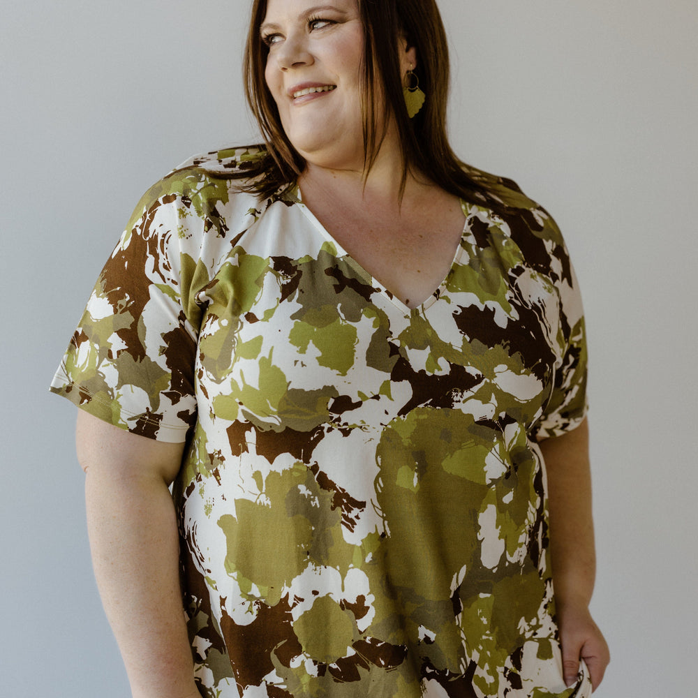 A person wearing the LARGE ABSTRACT FLORAL PRINT BLOUSE WITH RAGLAN SLEEVE by Yesta, featuring a green and brown pattern on soft jersey fabric, smiles as their earrings catch the light against a plain background.