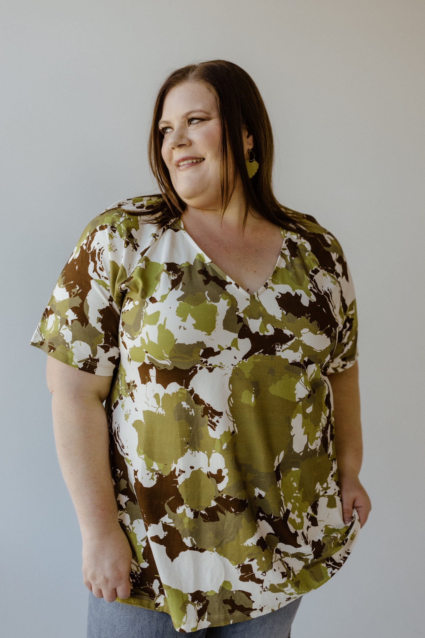 A person wearing the LARGE ABSTRACT FLORAL PRINT BLOUSE WITH RAGLAN SLEEVE by Yesta, featuring a green and brown pattern on soft jersey fabric, smiles as their earrings catch the light against a plain background.