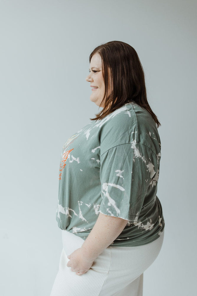 A person with long brown hair is facing away, wearing the "LET YOUR PROBLEMS FALL AWAY" graphic tee by Love Marlow, featuring a hand-bleached green tie-dye design, paired with a white skirt against a neutral background.