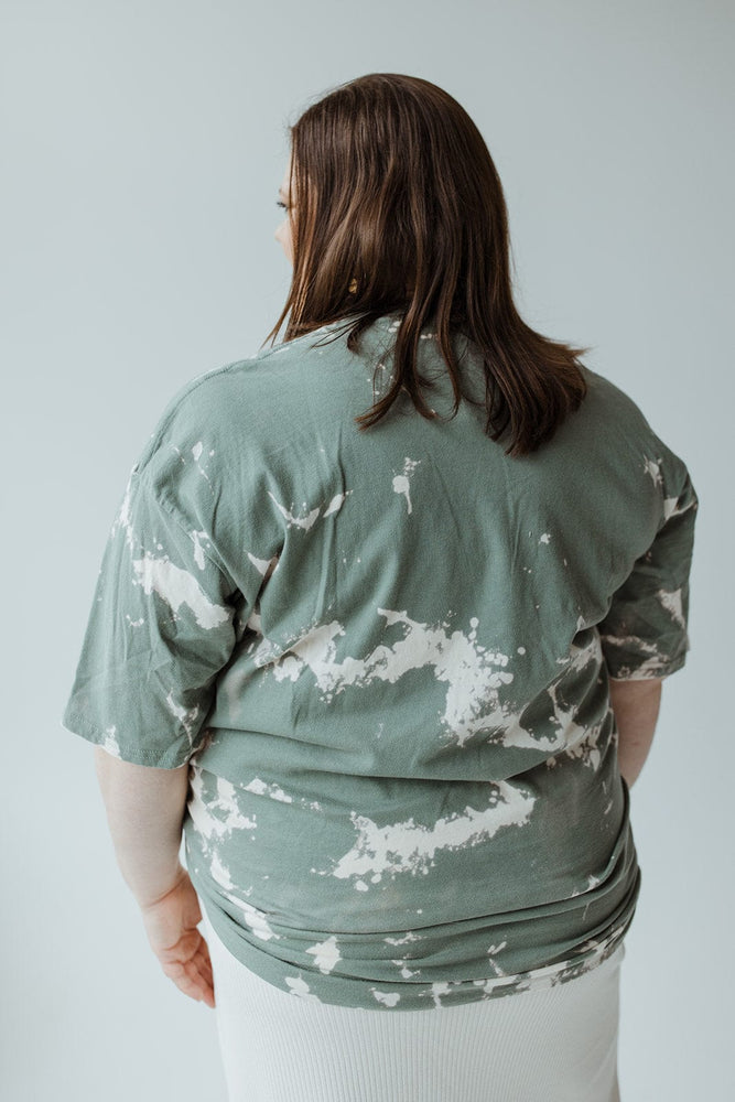 A person with long brown hair is facing away, wearing the "LET YOUR PROBLEMS FALL AWAY" graphic tee by Love Marlow, featuring a hand-bleached green tie-dye design, paired with a white skirt against a neutral background.