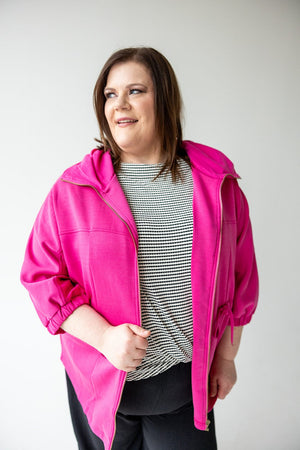 A woman wearing a bright pink LIGHT AS AIR CINCHED WAIST ZIP-UP by Love Marlow over a black and white striped top, standing and smiling.