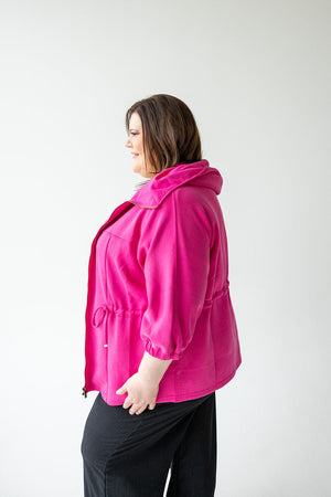 A person wearing a bright pink Love Marlow LIGHT AS AIR CINCHED WAIST ZIP-UP and black pants is standing and smiling in a side profile view against a plain white background.