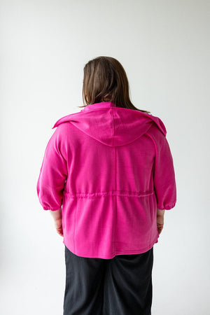 A person with long hair is standing with their back to the camera, wearing a bright pink Love Marlow LIGHT AS AIR CINCHED WAIST ZIP-UP and black pants, cinched at the waist, against a plain white background.