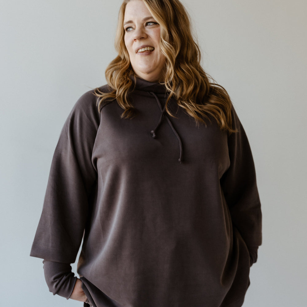 A person with long hair wearing a dark gray cowl neck tunic and pants smiles, looking to the side against a plain background, exuding an aura of luxurious comfort.