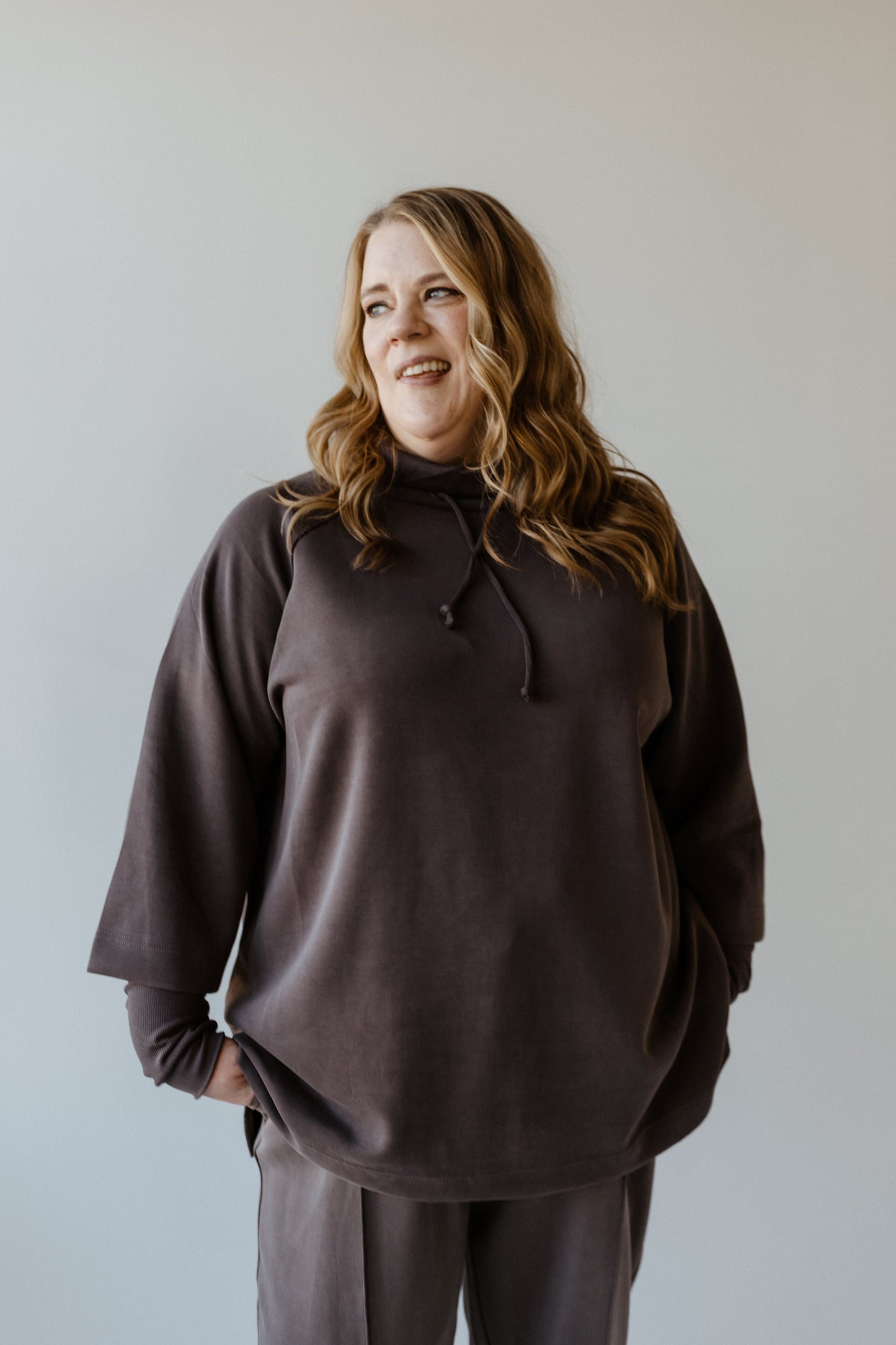 A person with long hair wearing a dark gray cowl neck tunic and pants smiles, looking to the side against a plain background, exuding an aura of luxurious comfort.