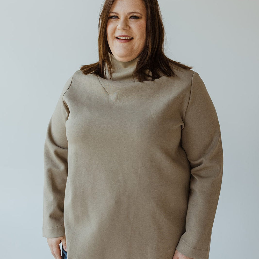 A woman with shoulder-length brown hair, wearing the sophisticated "LIGHT AS AIR DRAPE NECK IN SOFT ARMY" sweater by Yesta and a pair of jeans, stands against a plain background, smiling.