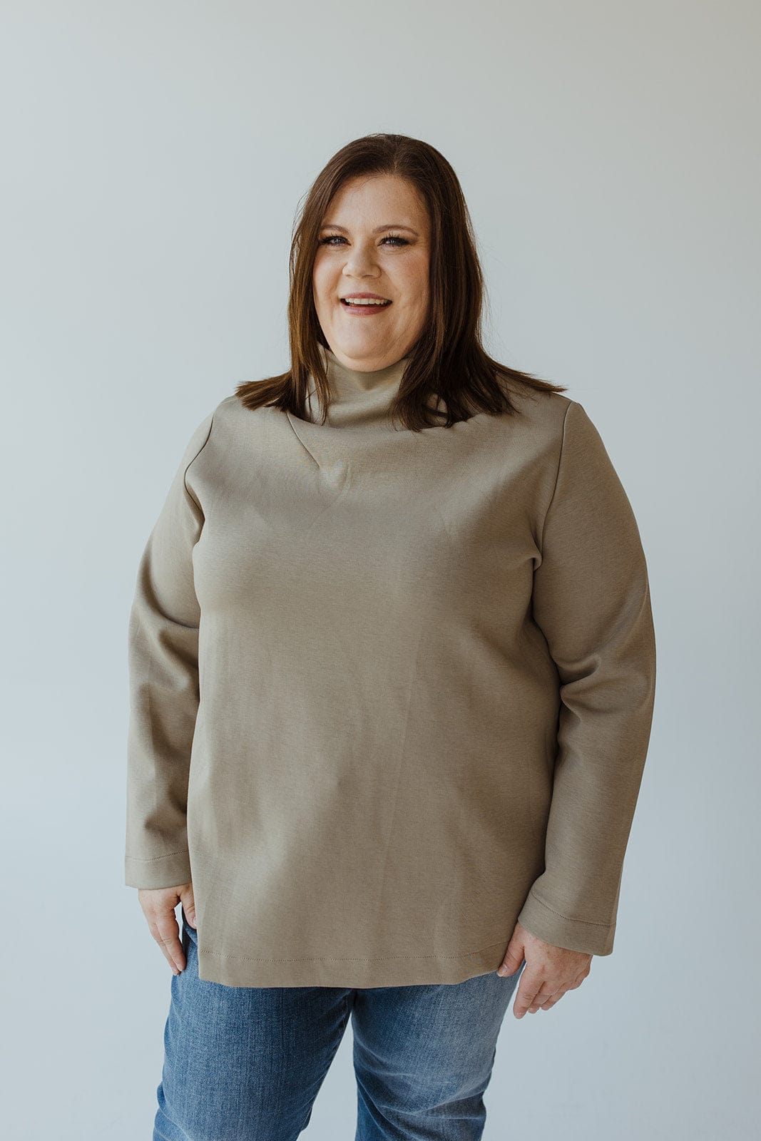 A woman with shoulder-length brown hair, wearing the sophisticated "LIGHT AS AIR DRAPE NECK IN SOFT ARMY" sweater by Yesta and a pair of jeans, stands against a plain background, smiling.