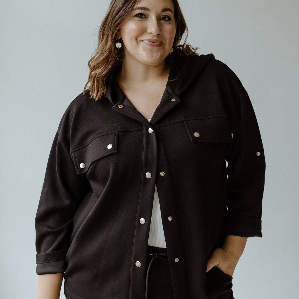 A person wearing a contemporary black jacket and earrings stands against a light gray background, smiling.