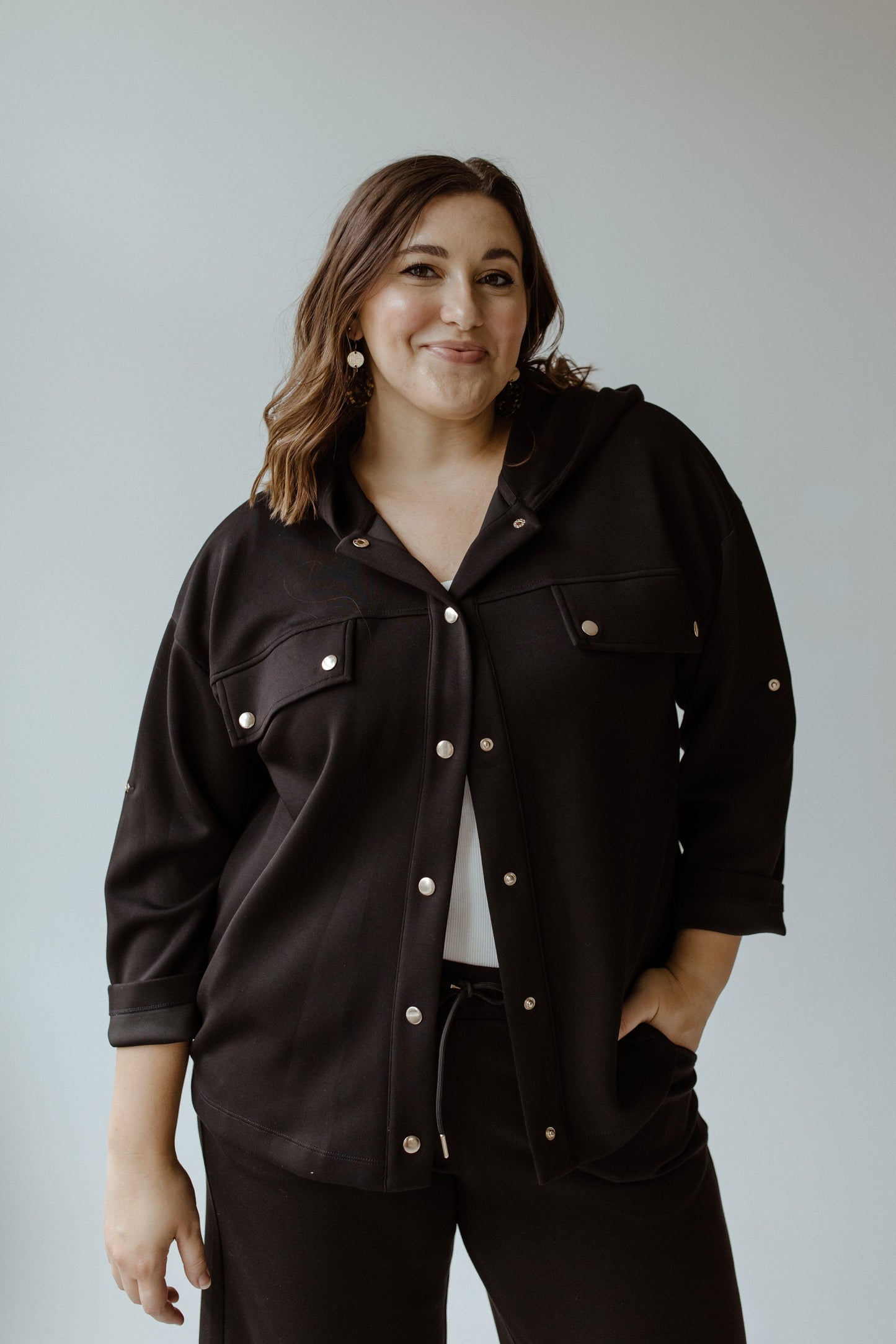 A person wearing a contemporary black jacket and earrings stands against a light gray background, smiling.