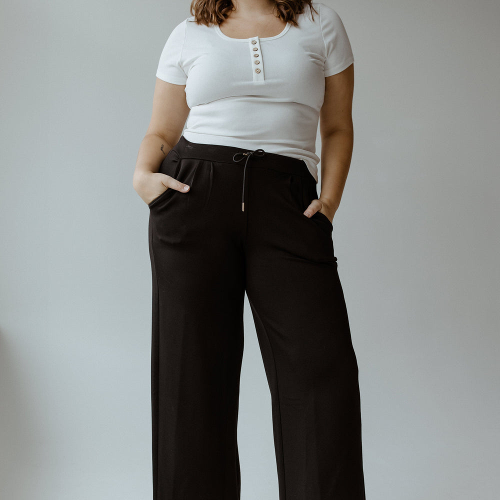 A person stands against a gray background, effortlessly capturing the athleisure vibe in a white t-shirt, Yesta's LIGHT AS AIR PLEATED WIDE LEG PANTS with an elastic waistband, and white sneakers. Hands tucked into pockets and a neutral expression complete the look.