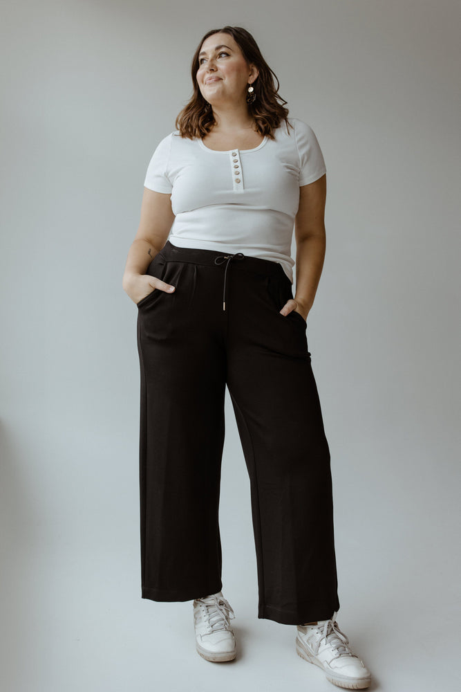 A person stands against a gray background, effortlessly capturing the athleisure vibe in a white t-shirt, Yesta's LIGHT AS AIR PLEATED WIDE LEG PANTS with an elastic waistband, and white sneakers. Hands tucked into pockets and a neutral expression complete the look.