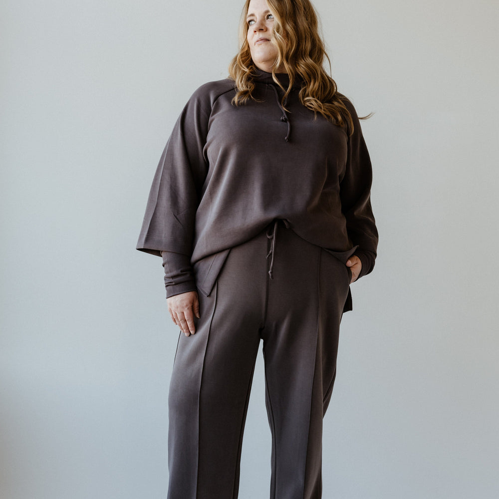 A person wearing a dark gray tracksuit, featuring relaxed slim straight pants and white shoes, stands in front of a plain light background. The attire gives an effortlessly stylish vibe, perfect for those who appreciate the comfort of loungewear that's as LIGHT AS AIR.