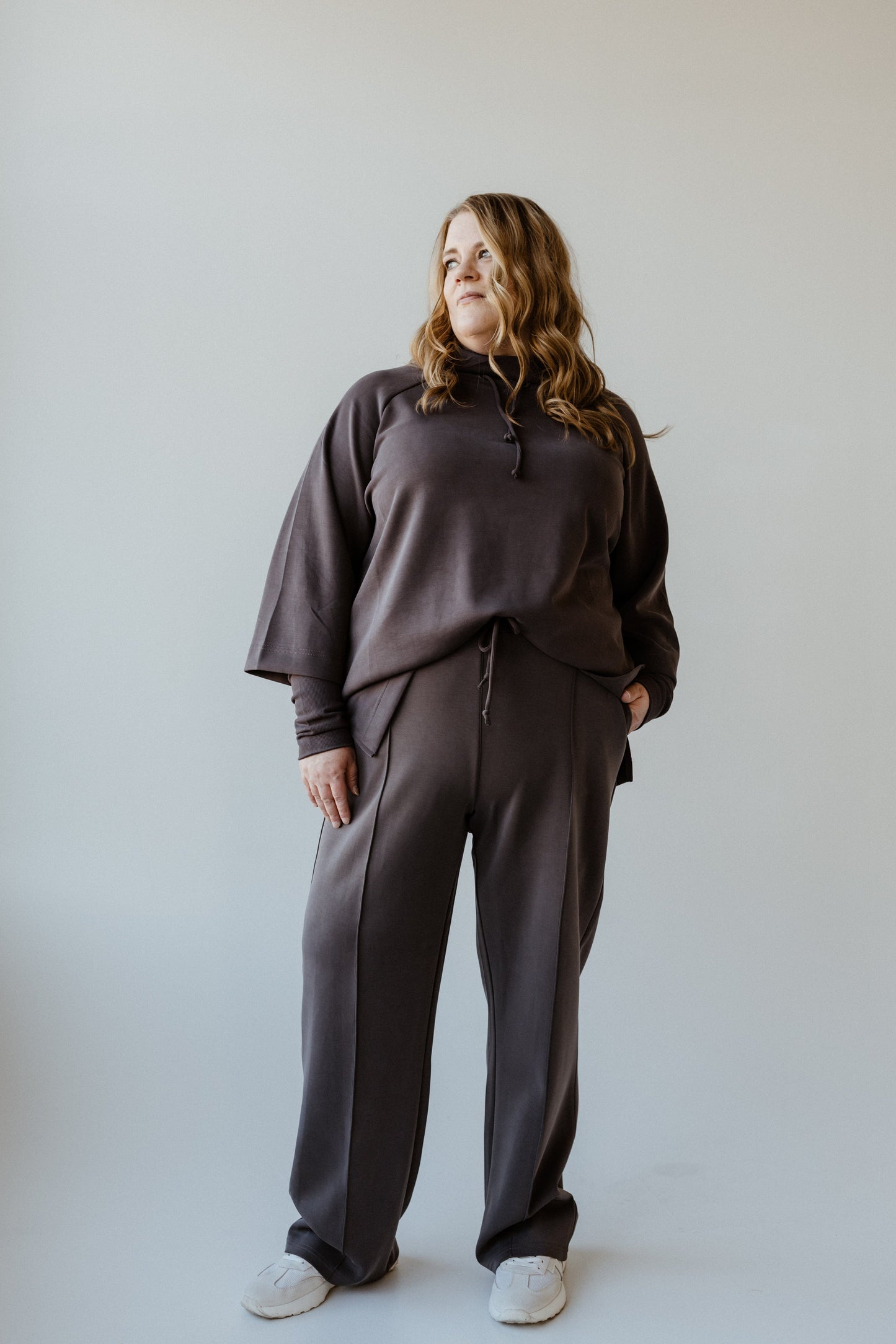 A person wearing a dark gray tracksuit, featuring relaxed slim straight pants and white shoes, stands in front of a plain light background. The attire gives an effortlessly stylish vibe, perfect for those who appreciate the comfort of loungewear that's as LIGHT AS AIR.