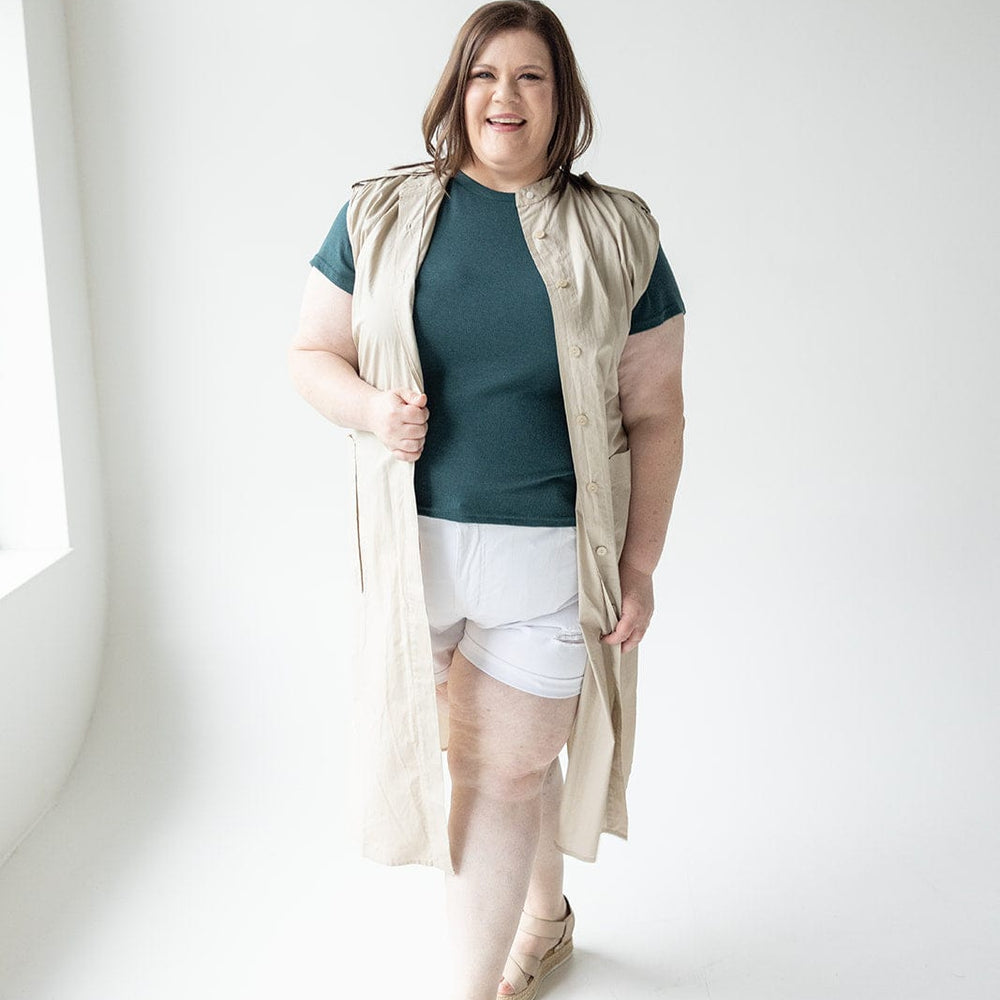 A person with shoulder-length hair wears a sleeveless beige Love Marlow LIGHTWEIGHT DUSTER VEST over a teal top and white shorts, standing and smiling in a well-lit room.