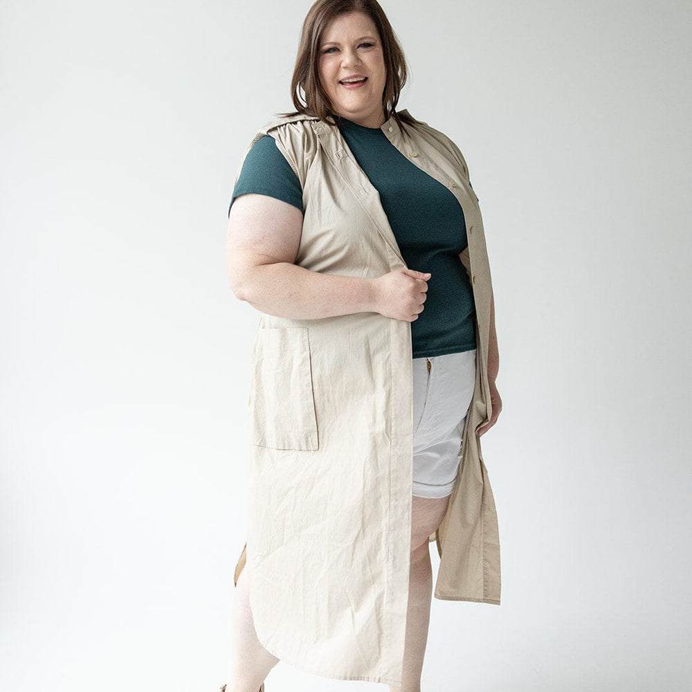 A person stands against a plain background, wearing a sleeveless beige Love Marlow LIGHTWEIGHT DUSTER VEST over a green shirt and white shorts, paired with beige sandals.