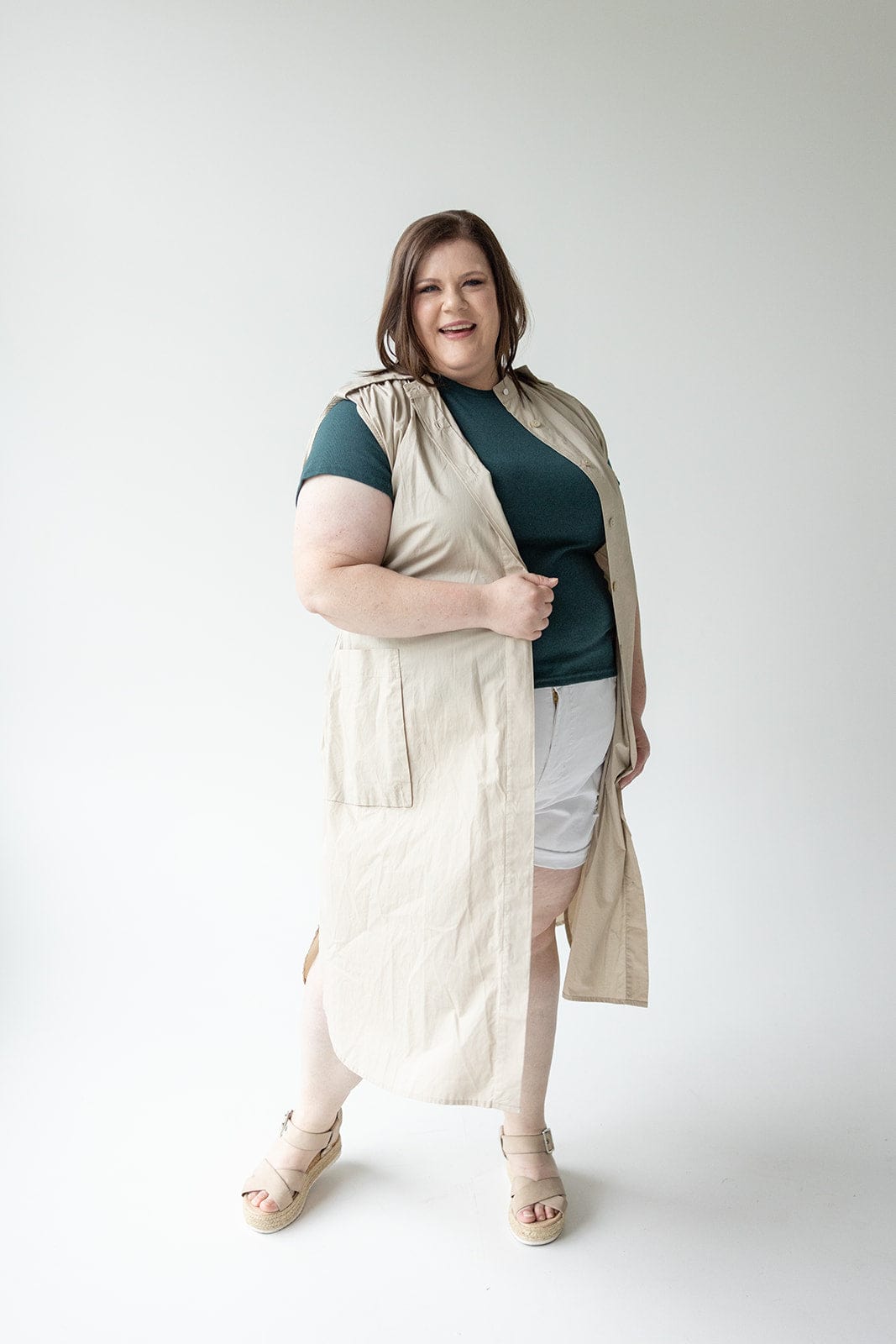 A person stands against a plain background, wearing a sleeveless beige Love Marlow LIGHTWEIGHT DUSTER VEST over a green shirt and white shorts, paired with beige sandals.