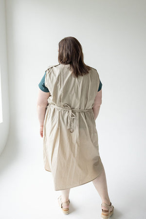 Person with shoulder-length brown hair wearing a sleeveless beige tunic dress tied at the waist and a Love Marlow LIGHTWEIGHT DUSTER VEST, standing with back to the camera in a white, well-lit room.