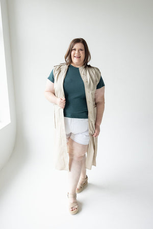 A person with shoulder-length hair wears a sleeveless beige Love Marlow LIGHTWEIGHT DUSTER VEST over a teal top and white shorts, standing and smiling in a well-lit room.