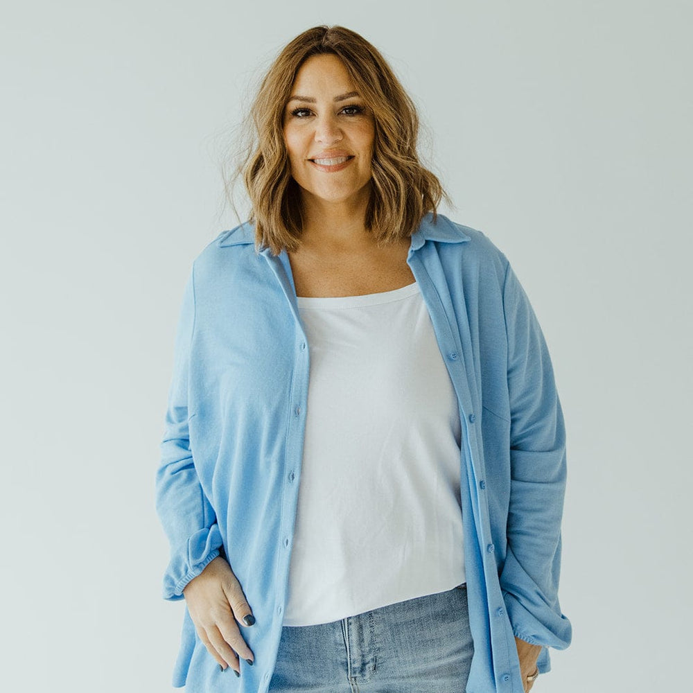 A person with shoulder-length hair stands against a plain background, donning an understated luxury ensemble: the Love Marlow LIGHTWEIGHT KNIT BUTTON-DOWN IN AZURE over a white top and blue jeans.