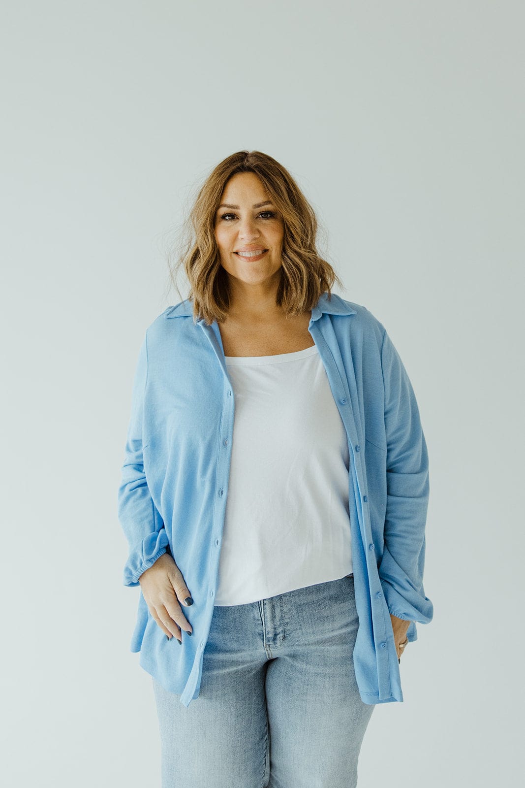 A person with shoulder-length hair stands against a plain background, donning an understated luxury ensemble: the Love Marlow LIGHTWEIGHT KNIT BUTTON-DOWN IN AZURE over a white top and blue jeans.