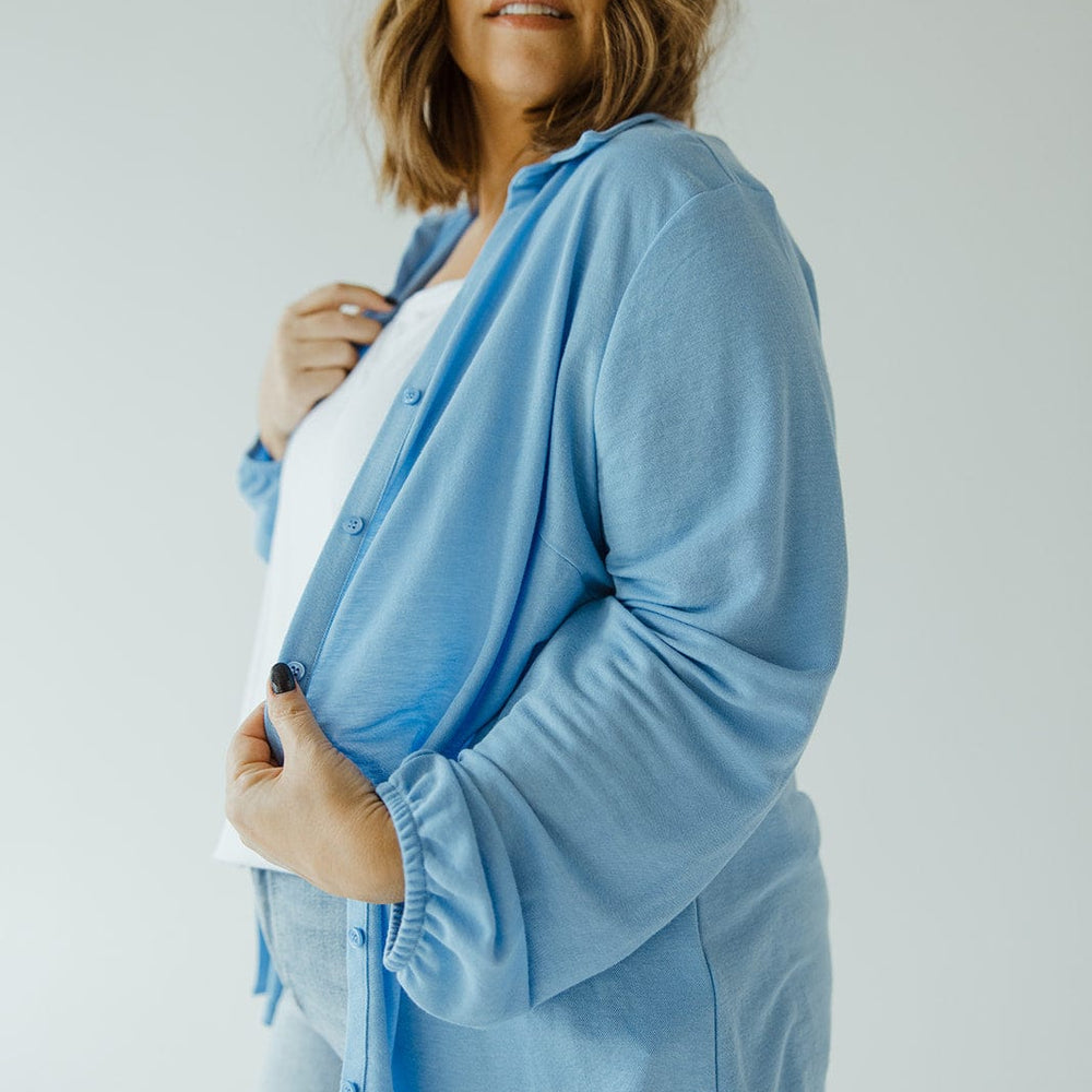 A woman with shoulder-length hair, dressed in Love Marlow's understated luxury LIGHTWEIGHT KNIT BUTTON-DOWN IN AZURE, a white shirt, and light blue jeans, stands against a plain background.