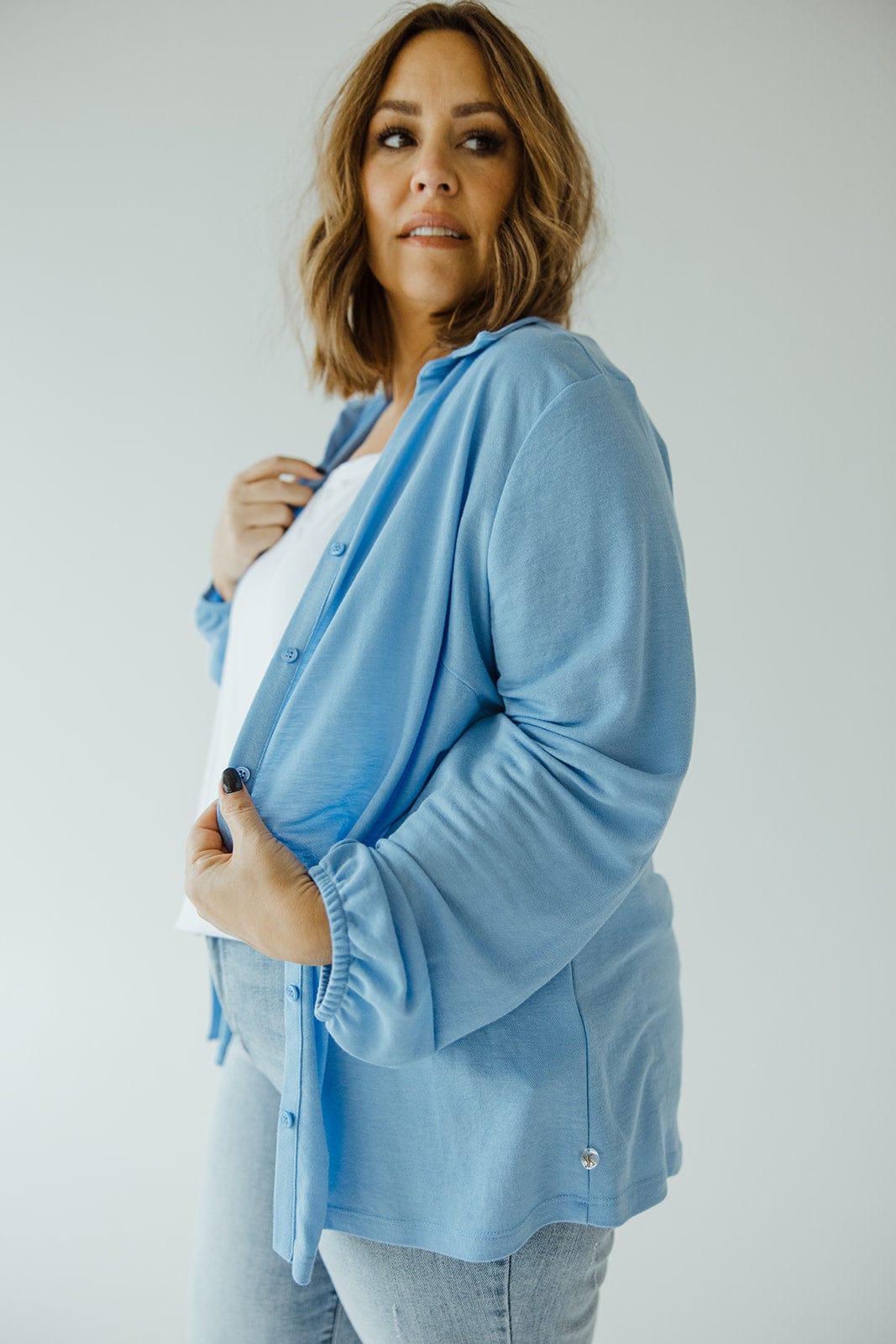 A woman with shoulder-length hair, dressed in Love Marlow's understated luxury LIGHTWEIGHT KNIT BUTTON-DOWN IN AZURE, a white shirt, and light blue jeans, stands against a plain background.