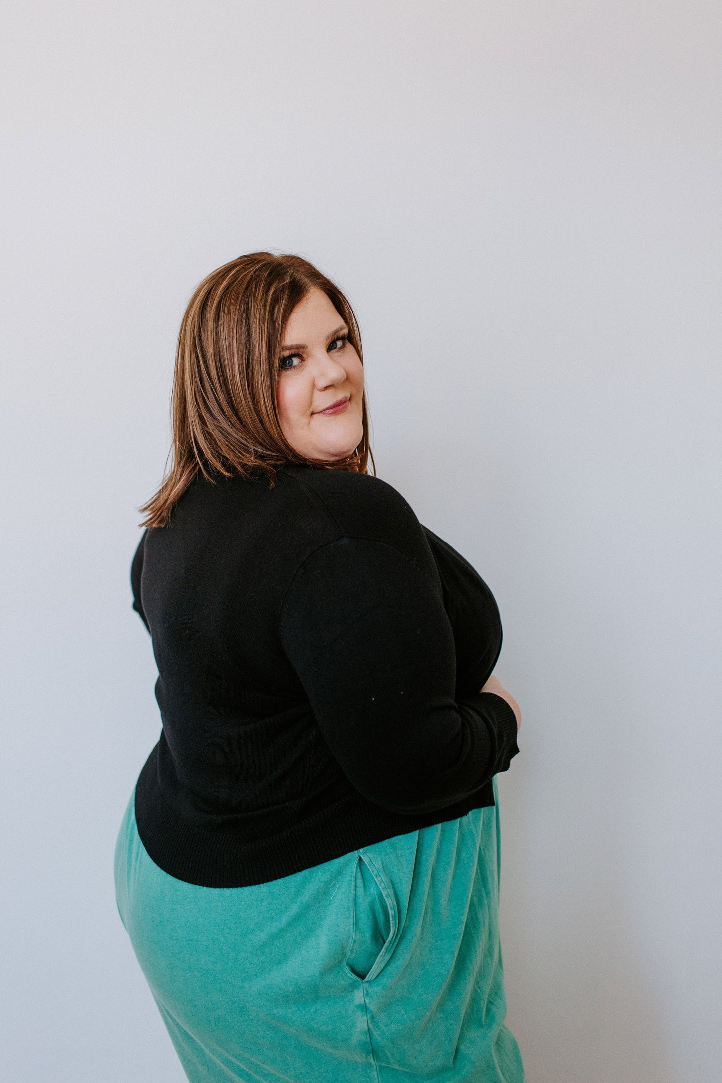 A woman wearing a black top and turquoise skirt stands against a plain white background. The snug fit accentuates her silhouette as she looks over her shoulder at the camera, creating an effortlessly chic look.