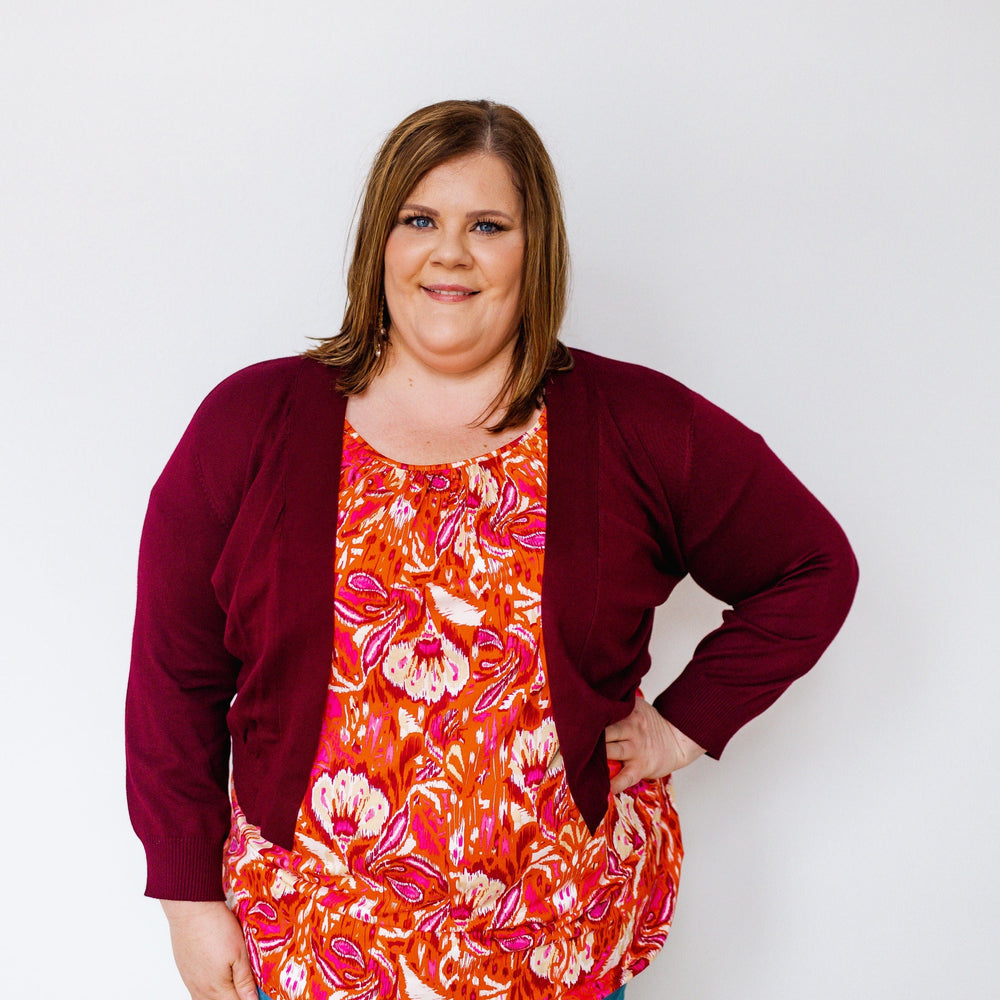 A person wearing a burgundy cardigan, a vibrant floral top, and blue jeans stands against a plain white background.