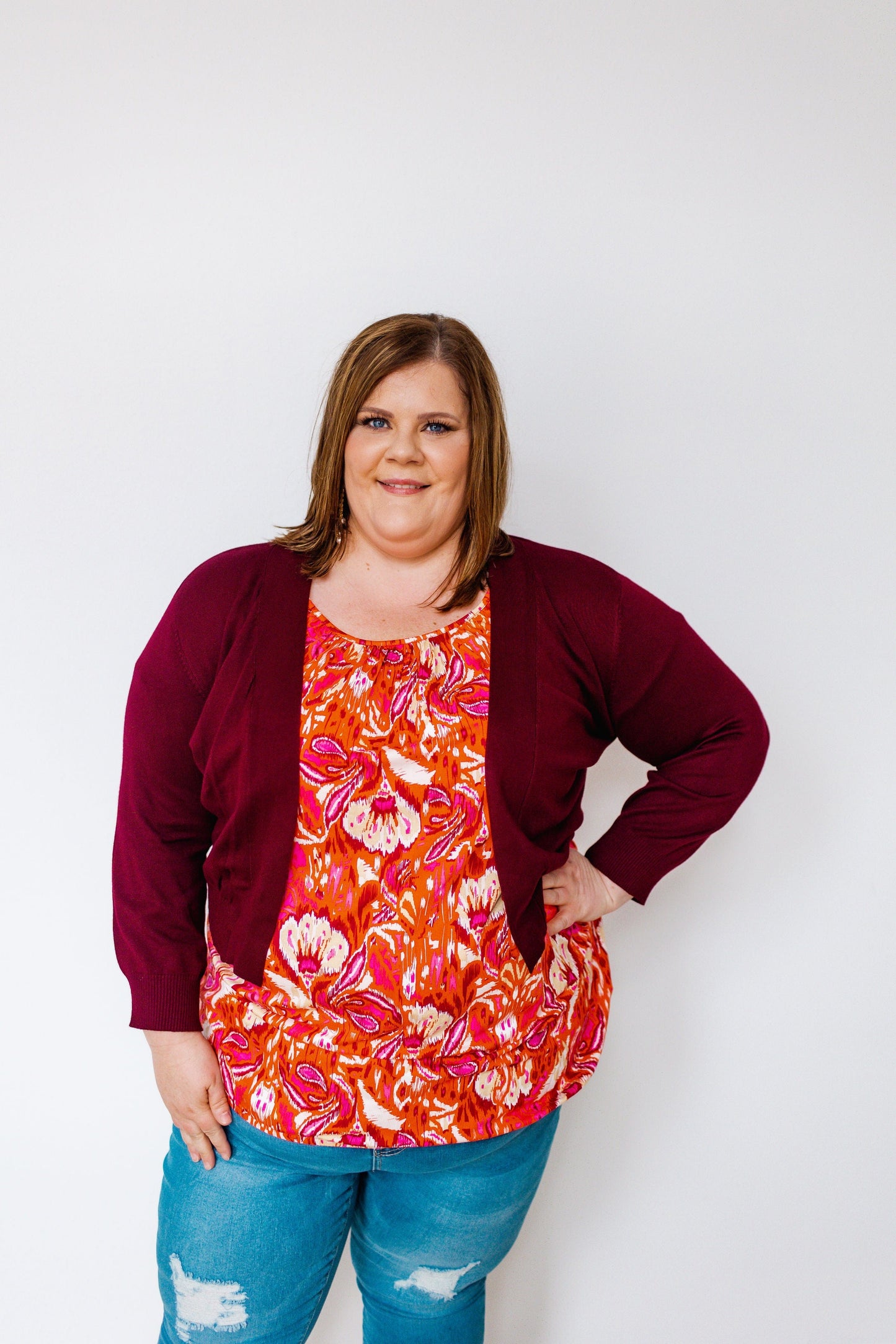 A person wearing a burgundy cardigan, a vibrant floral top, and blue jeans stands against a plain white background.