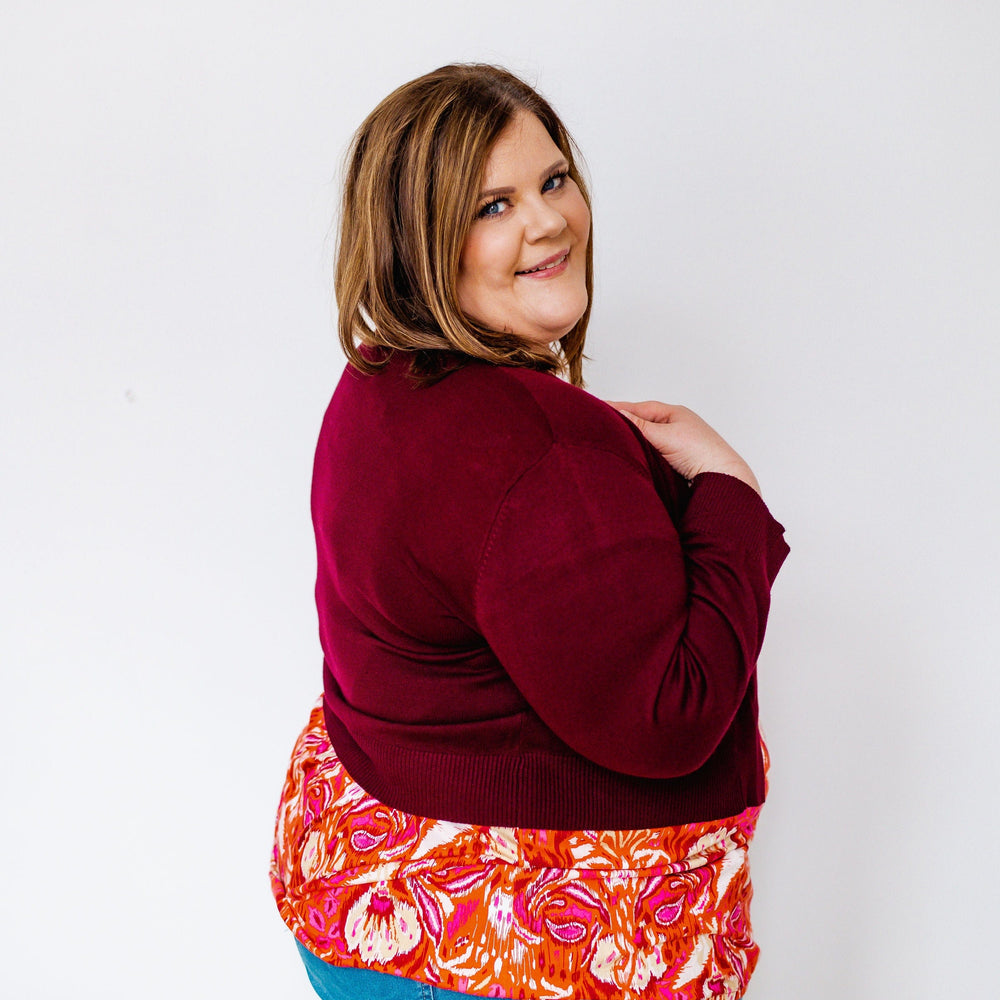 A person in a burgundy floral-patterned top and jeans poses gracefully against a plain white background, adding a touch of charm with a lightweight cardigan draped over their shoulders.