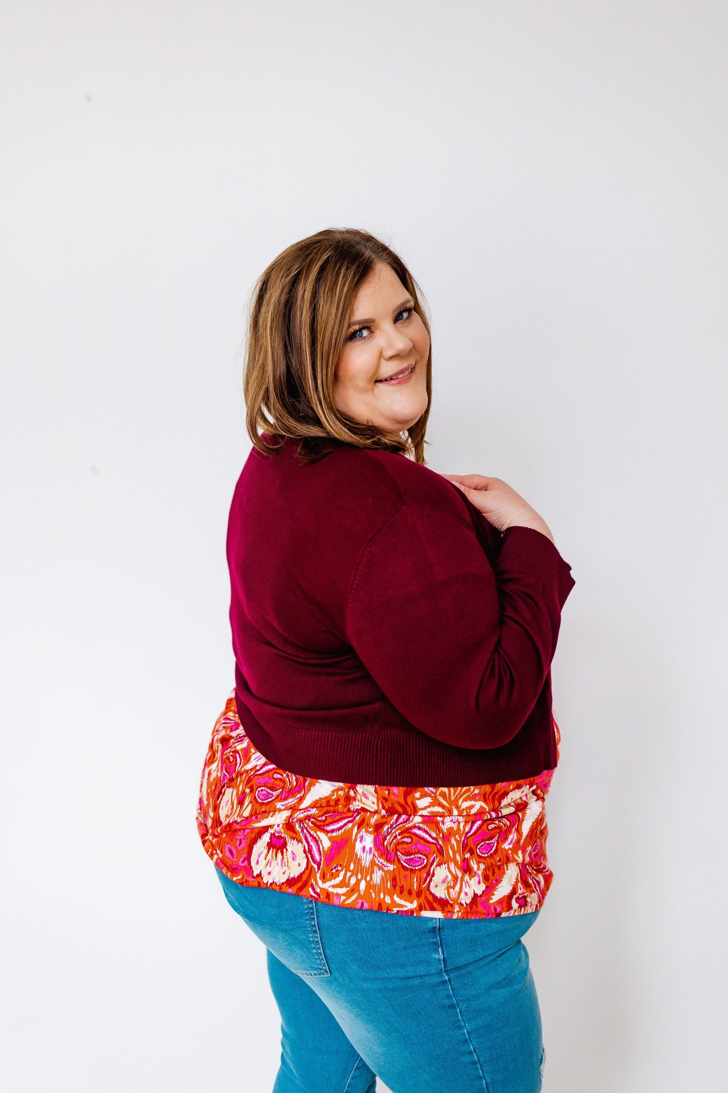 A person in a burgundy floral-patterned top and jeans poses gracefully against a plain white background, adding a touch of charm with a lightweight cardigan draped over their shoulders.