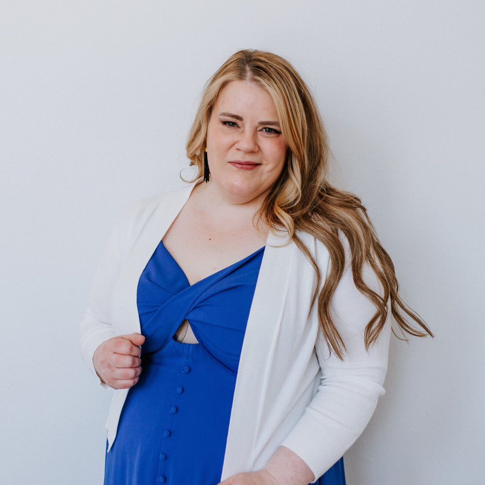 A woman with long hair wearing a blue dress and a lightweight cardigan with 3/4 sleeves stands against a plain white background.