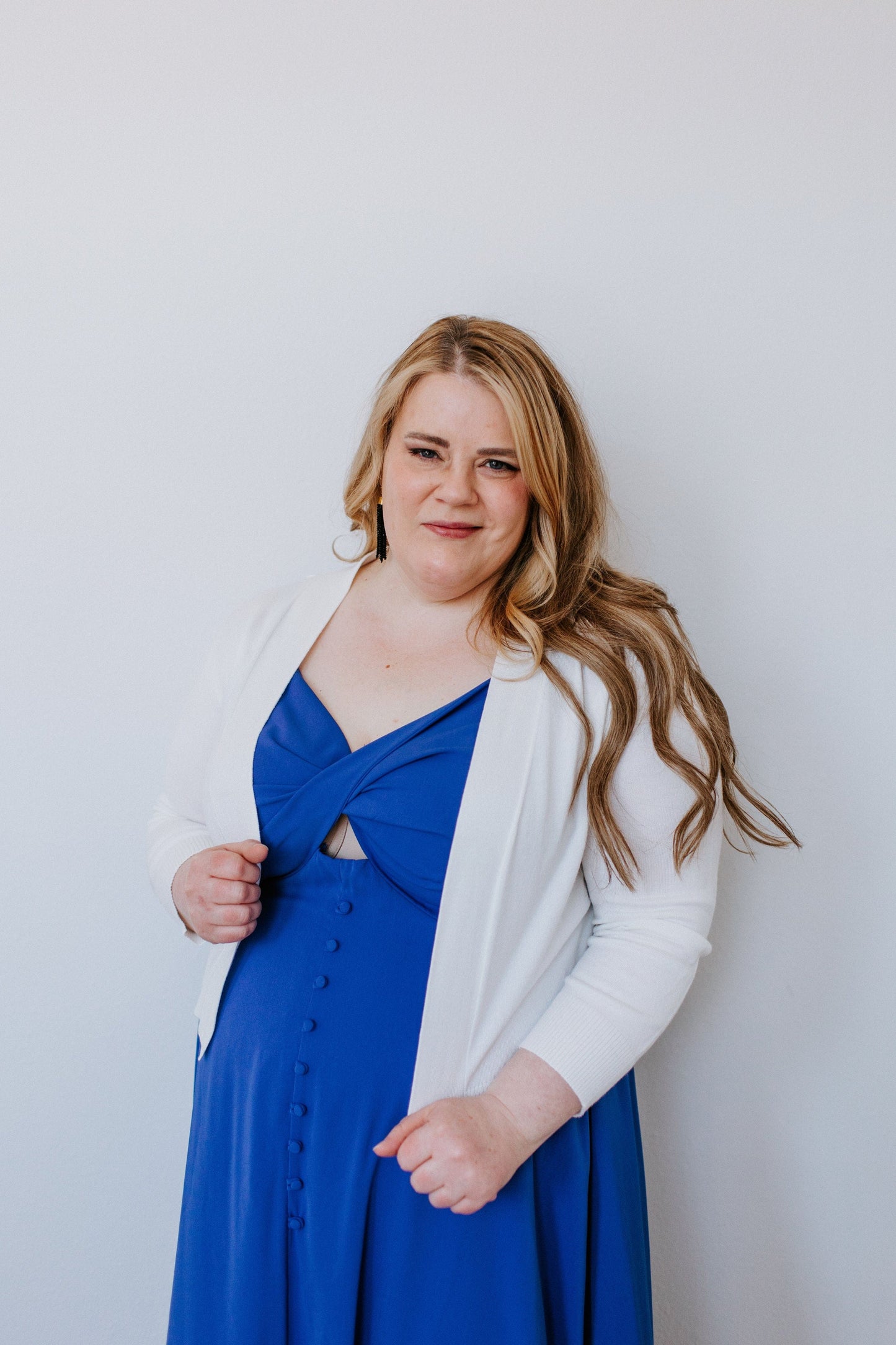 A woman with long hair wearing a blue dress and a lightweight cardigan with 3/4 sleeves stands against a plain white background.