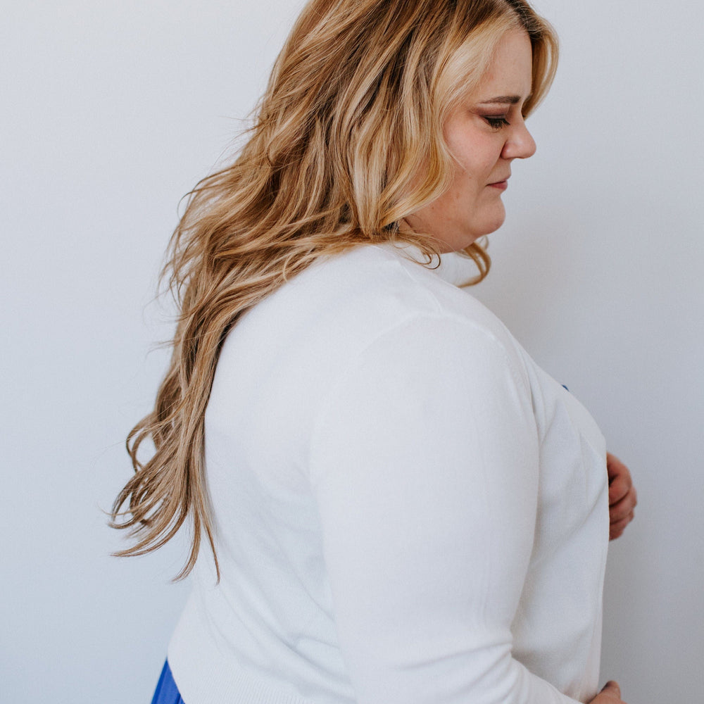 A person with long hair is wearing a white sweater with 3/4 sleeves and a blue skirt, standing sideways against a plain white background.