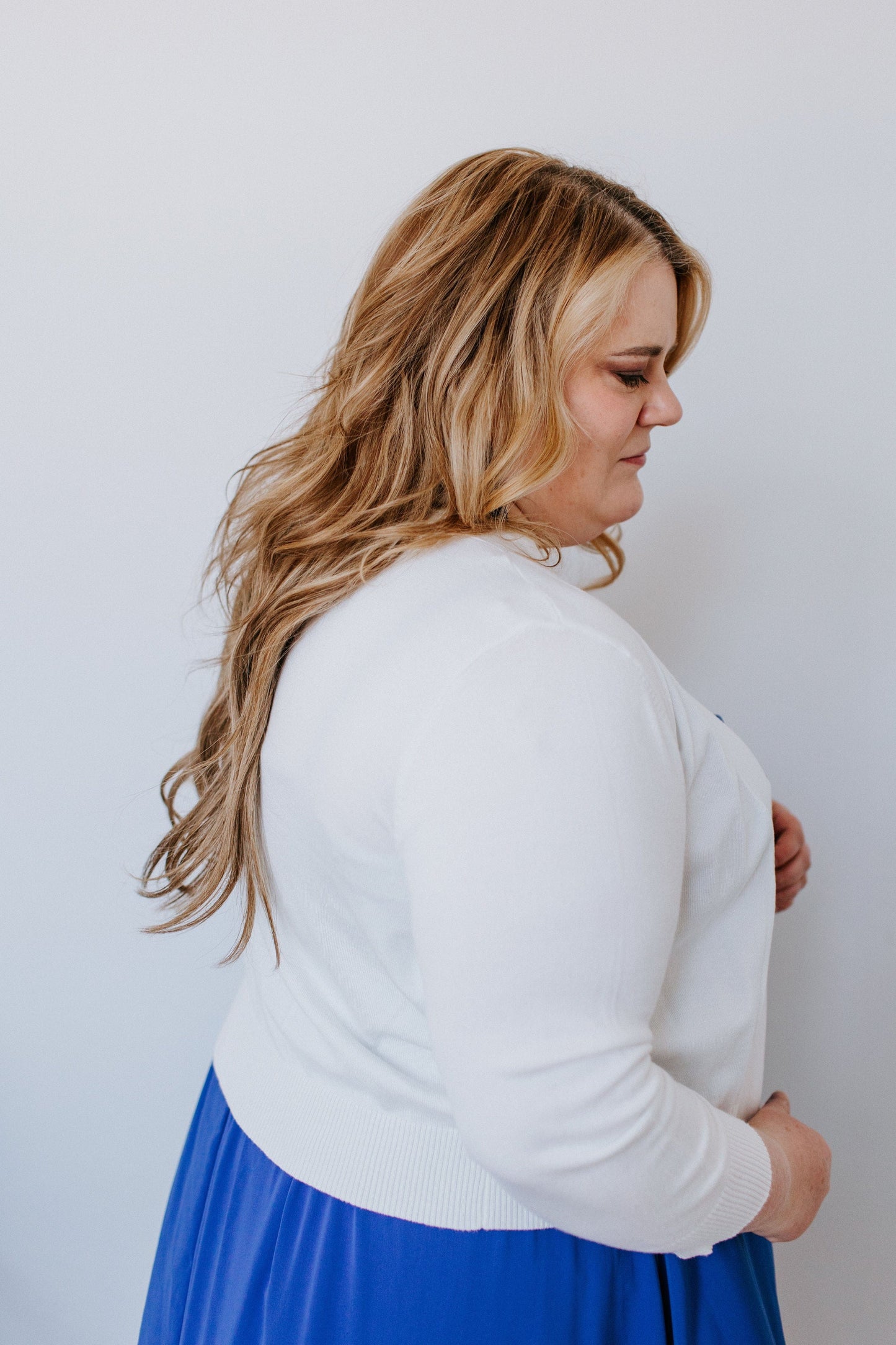 A person with long hair is wearing a white sweater with 3/4 sleeves and a blue skirt, standing sideways against a plain white background.