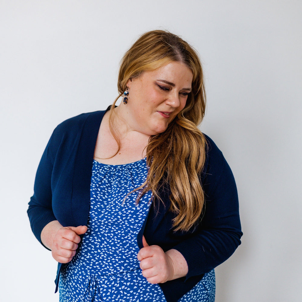 A person with long hair wearing a blue patterned dress and a navy cardigan with 3/4 length sleeves looks down while standing against a plain background.