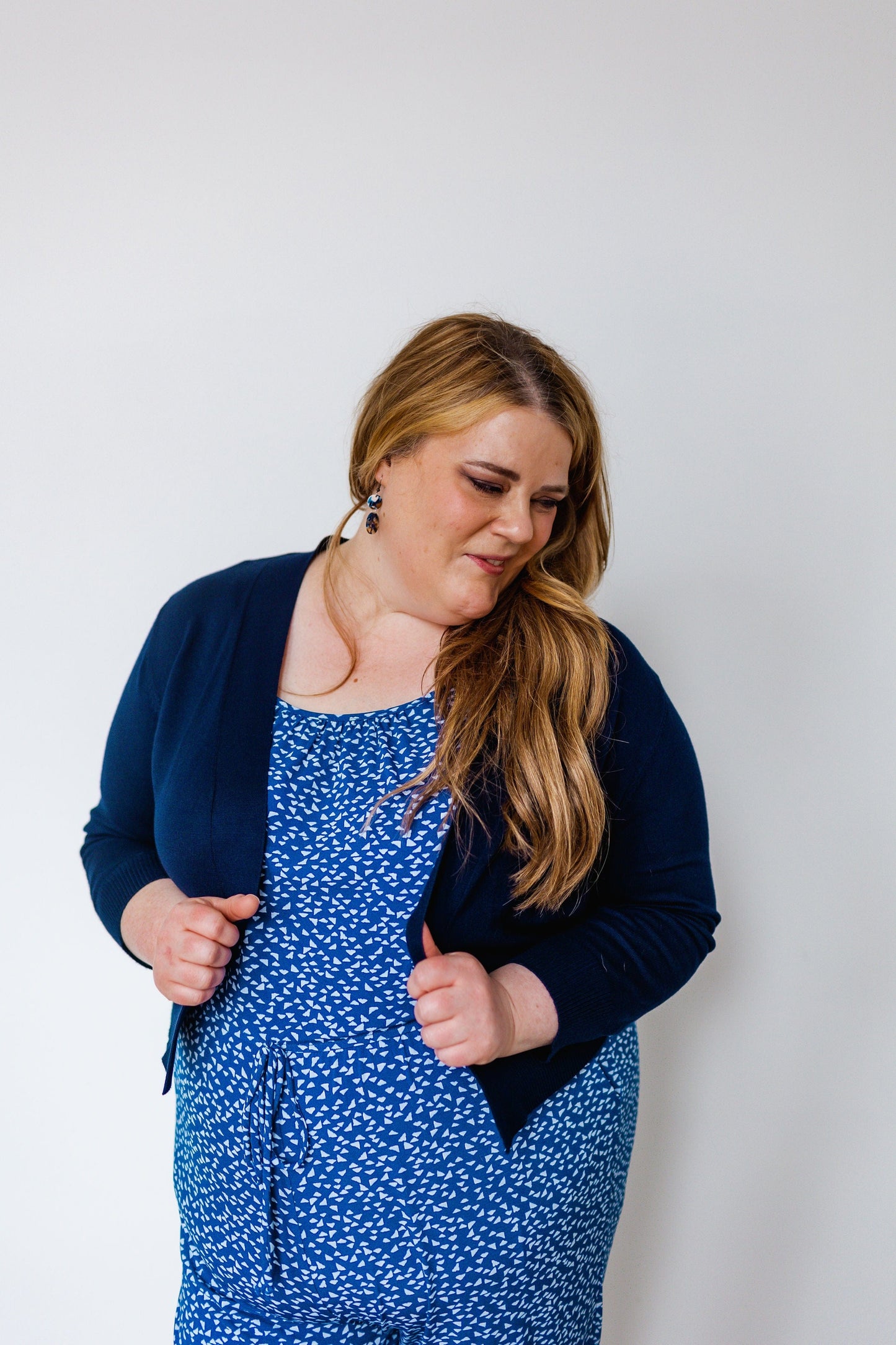 A person with long hair wearing a blue patterned dress and a navy cardigan with 3/4 length sleeves looks down while standing against a plain background.