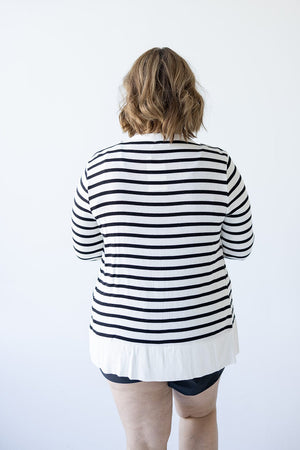 A person with shoulder-length hair, shown from the back, stands against a plain white background wearing black shorts and the Love Marlow Lightweight Striped Snap Cardigan in Ivory and Black.