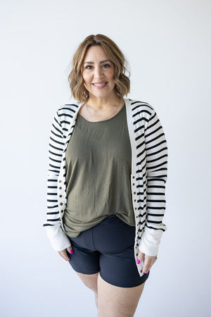 A woman with wavy hair stands against a plain white background, wearing Love Marlow's Lightweight Striped Snap Cardigan in Ivory and Black over an olive green shirt and black shorts, exuding chic comfort with her hands in her pockets.