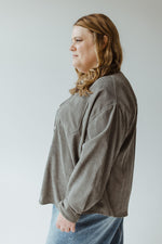 A person with long, wavy, light brown hair is seen from behind, wearing the Lightweight Corduroy Jacket in Frosted Olive by Mikarose and light blue jeans. The background is plain and light-colored.