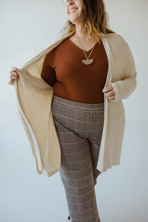 A person in a brown top, plaid pants, and a Yesta Lightweight Long-Sleeved Duster Cardigan in classic sand is smiling while holding the sides of the cardigan open.