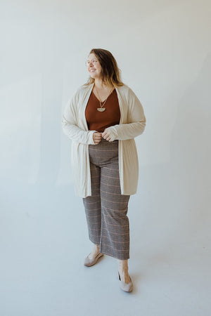 A person with glasses stands smiling, wearing Yesta's Lightweight Long-Sleeved Duster Cardigan in Sand over a brown top and checkered pants with beige shoes, against a plain white background.