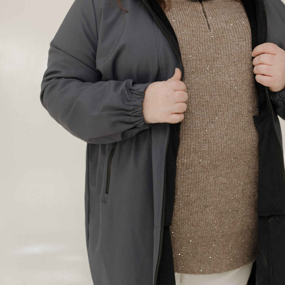 Person wearing a gray coat over a brown sweater and white pants on a plain background
