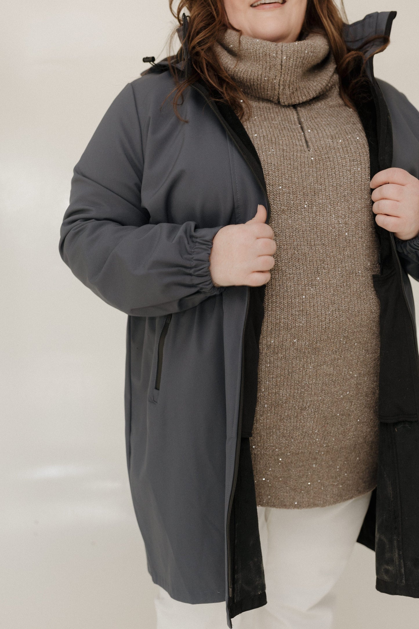 Person wearing a gray coat over a brown sweater and white pants on a plain background