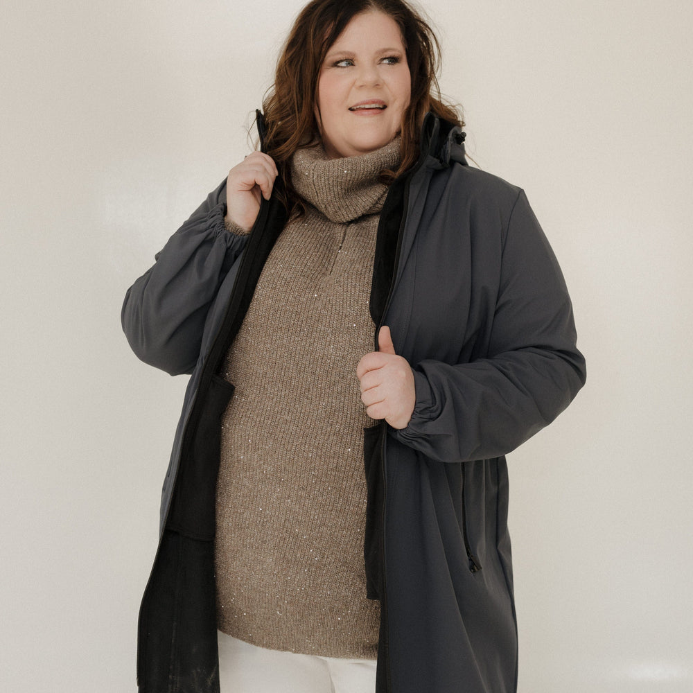 Woman wearing a dark gray coat over a brown sweater and white pants on a plain background