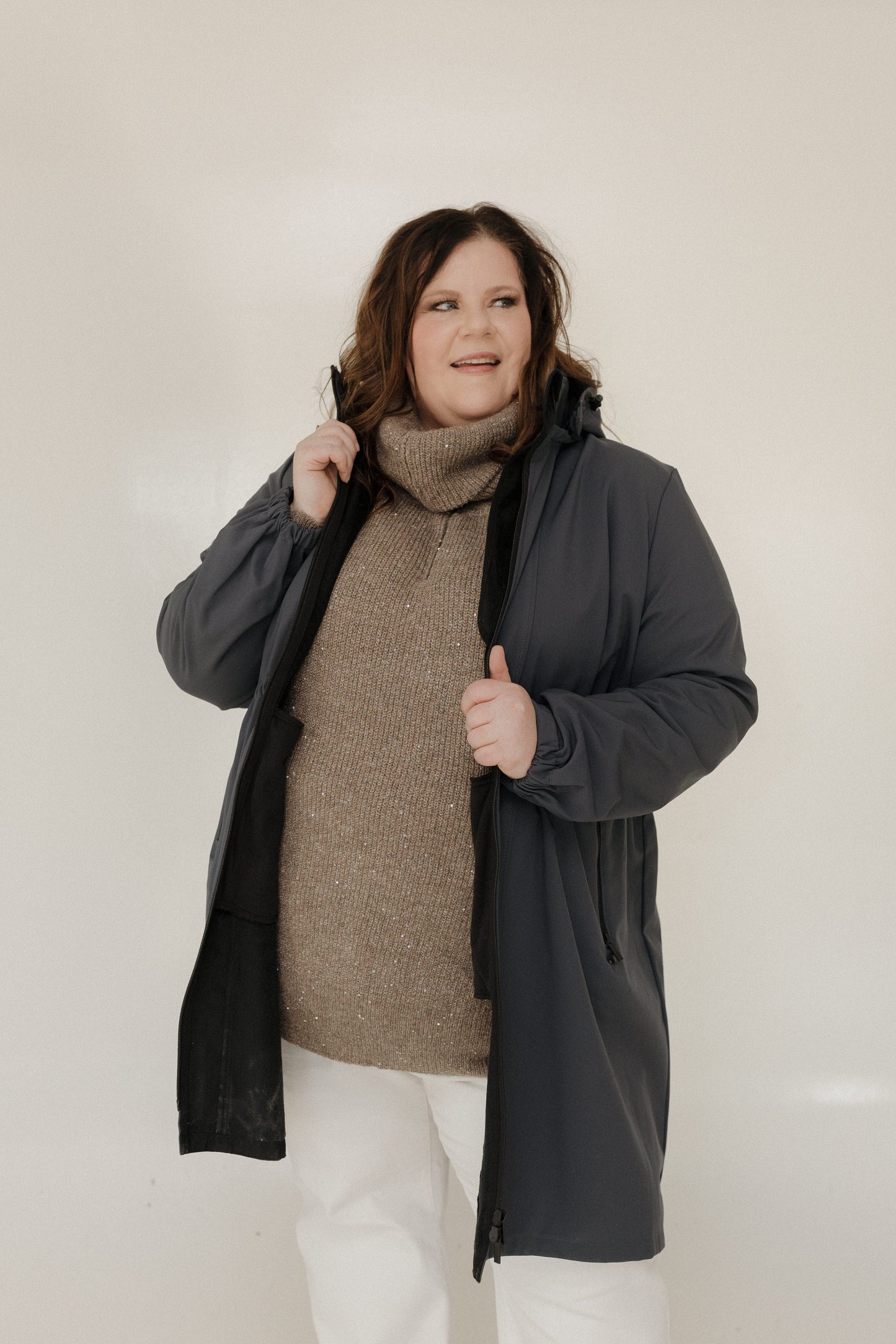 Woman wearing a dark gray coat over a brown sweater and white pants on a plain background