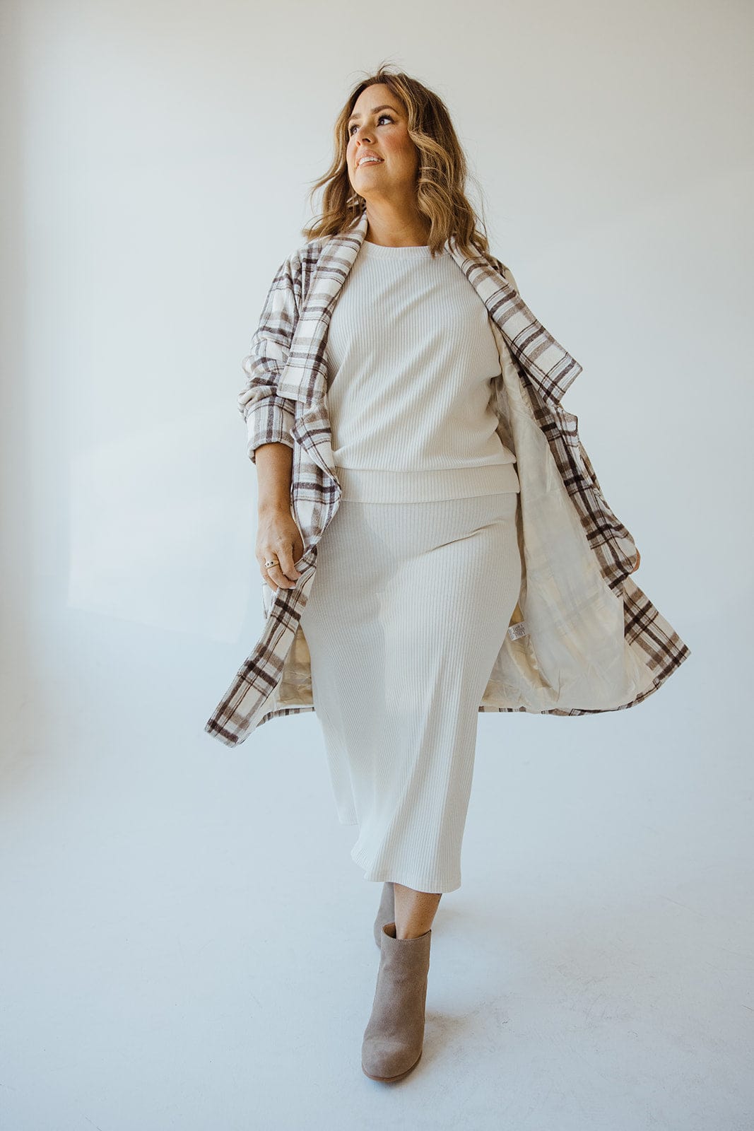 An individual stands confidently in a white outfit, wearing the LIGHTWEIGHT PLAID MIDI BLAZER by Andrea by Unit. They look upwards, showcasing brown ankle boots in a well-lit room with a white background.